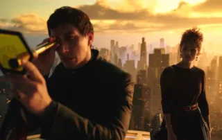zwei Menschen stehen auf dem Dach eines Wolkenkratzers, im Hintergrund ragen die Häuser einer Stadt im dämmernden Sonnenlicht nach oben, links im Bild steht ein Mann in dunkler Kleidung, der durch ein fernglasähnliches Gerät blickt, hinter ihm steht eine Frau, die ihn mustert