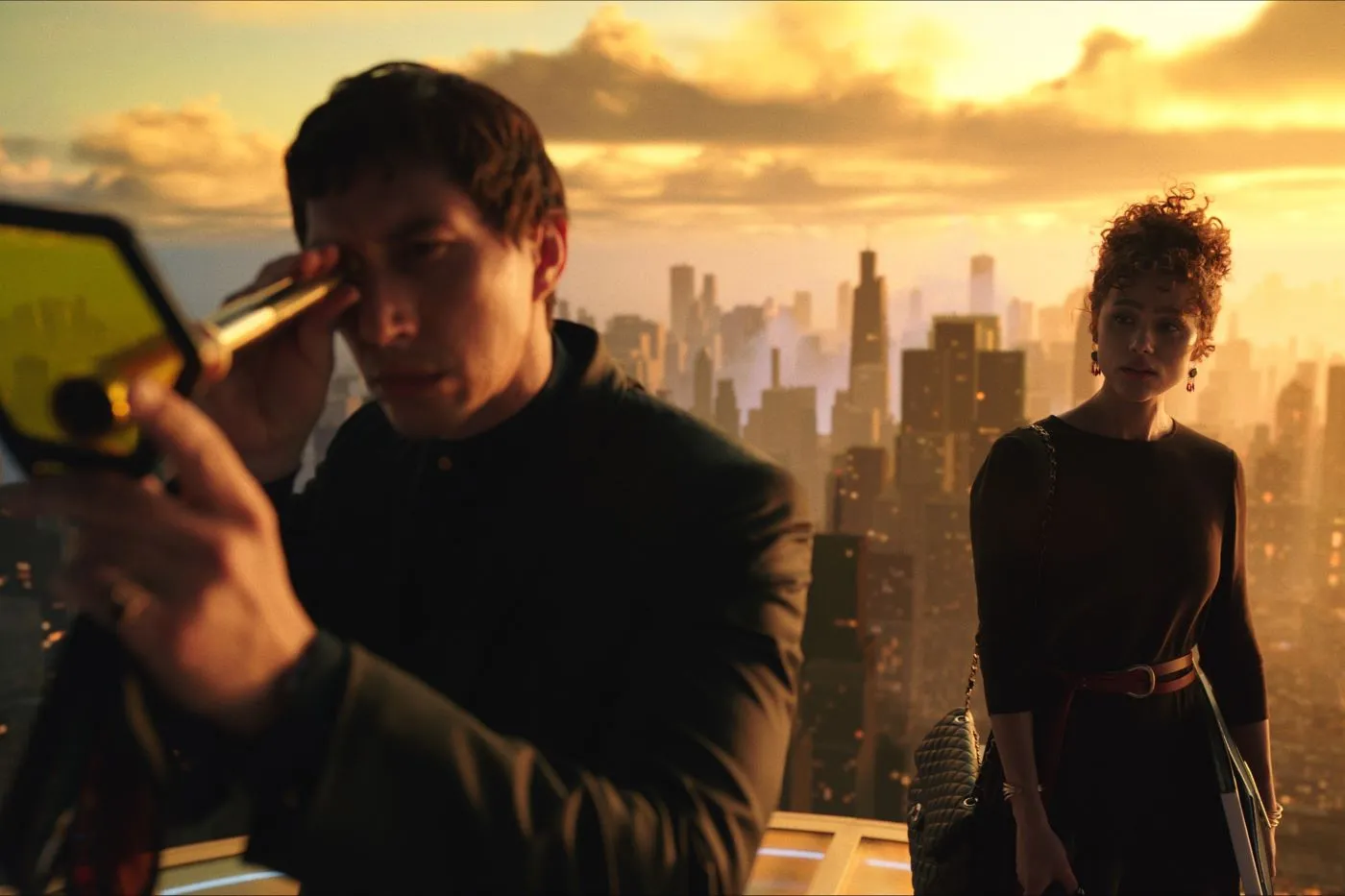 zwei Menschen stehen auf dem Dach eines Wolkenkratzers, im Hintergrund ragen die Häuser einer Stadt im dämmernden Sonnenlicht nach oben, links im Bild steht ein Mann in dunkler Kleidung, der durch ein fernglasähnliches Gerät blickt, hinter ihm steht eine Frau, die ihn mustert