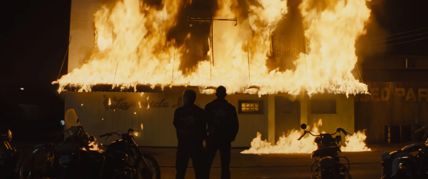 Nacht, aus einem Gebäude schlagen hohe Flammen, vor dem brennenden Haus stehen zwei Menschen mit dem Rücken zur Kamera, links und rechts von ihnen stehen Motorräder