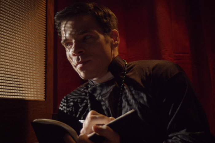 ein dunkel gekleideter Priester mit einem Notizbuch in der Hand sitzt auf dem Beichtstuhl