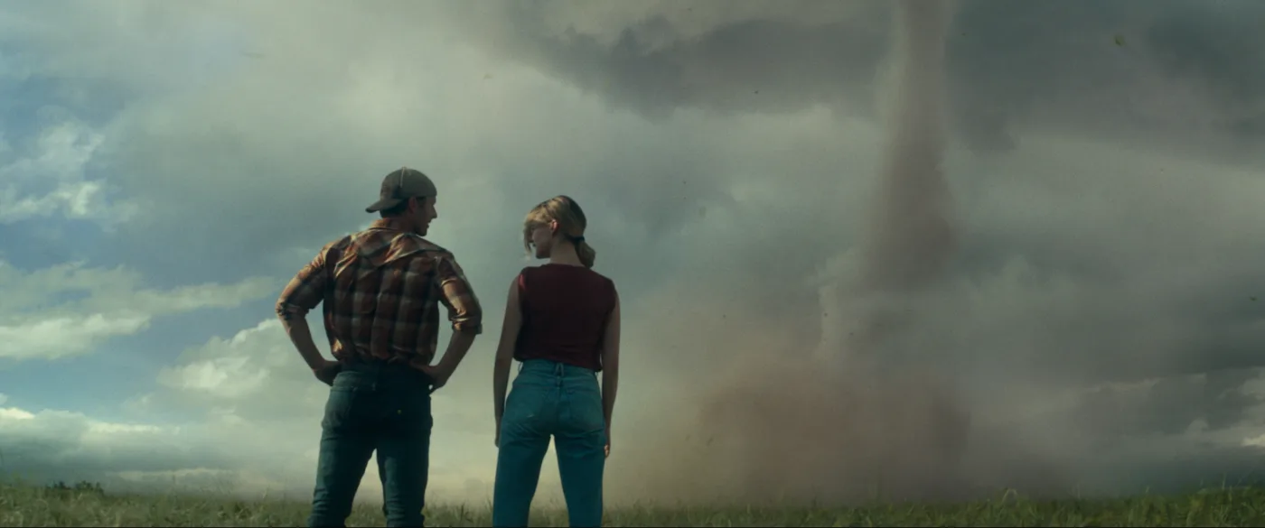 ein Mann im Hemd, langer Hose und Cap und eine Frau im T-Shirt und Jeans stehen nebeneinander mit dem Rücken zur Kamera, vor ihnen baut sich auf flacher Landschaft ein Tornado auf