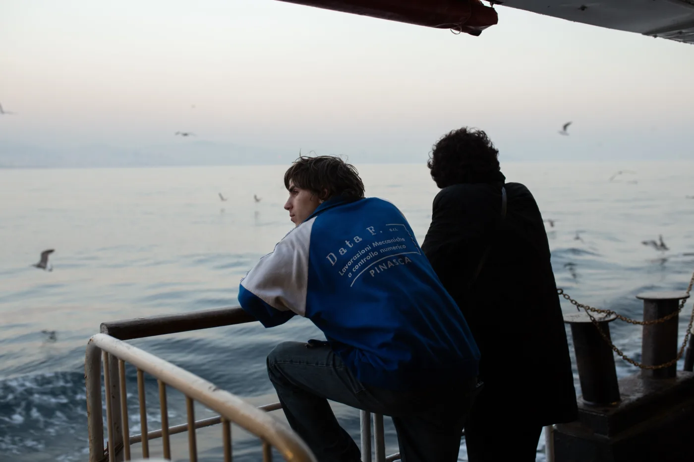 ein junger Mann in blau-weißer Jacke und eine Frau im schwarzen Mantel stehen mit dem Rücken zur Kamera an der Reling eines Schiffes, sie sehen hinaus aufs Meer, Möwen fliegen umher