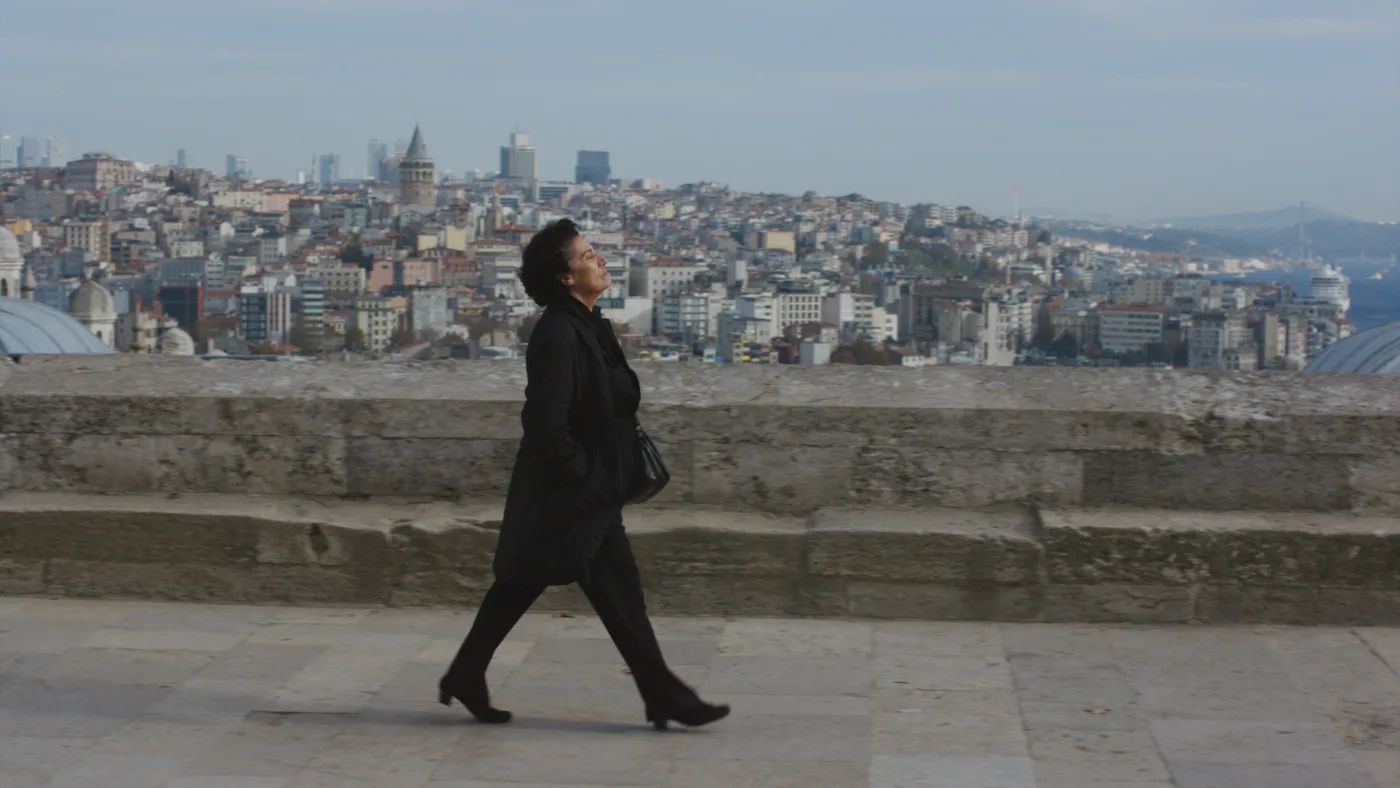 ein Frau im schwarzen Mantel, schwarzer Hose und schwarzen Schuhen geht einen Bürgersteig entlang, im Hintergrund baut sich das Panorama einer Stadt auf