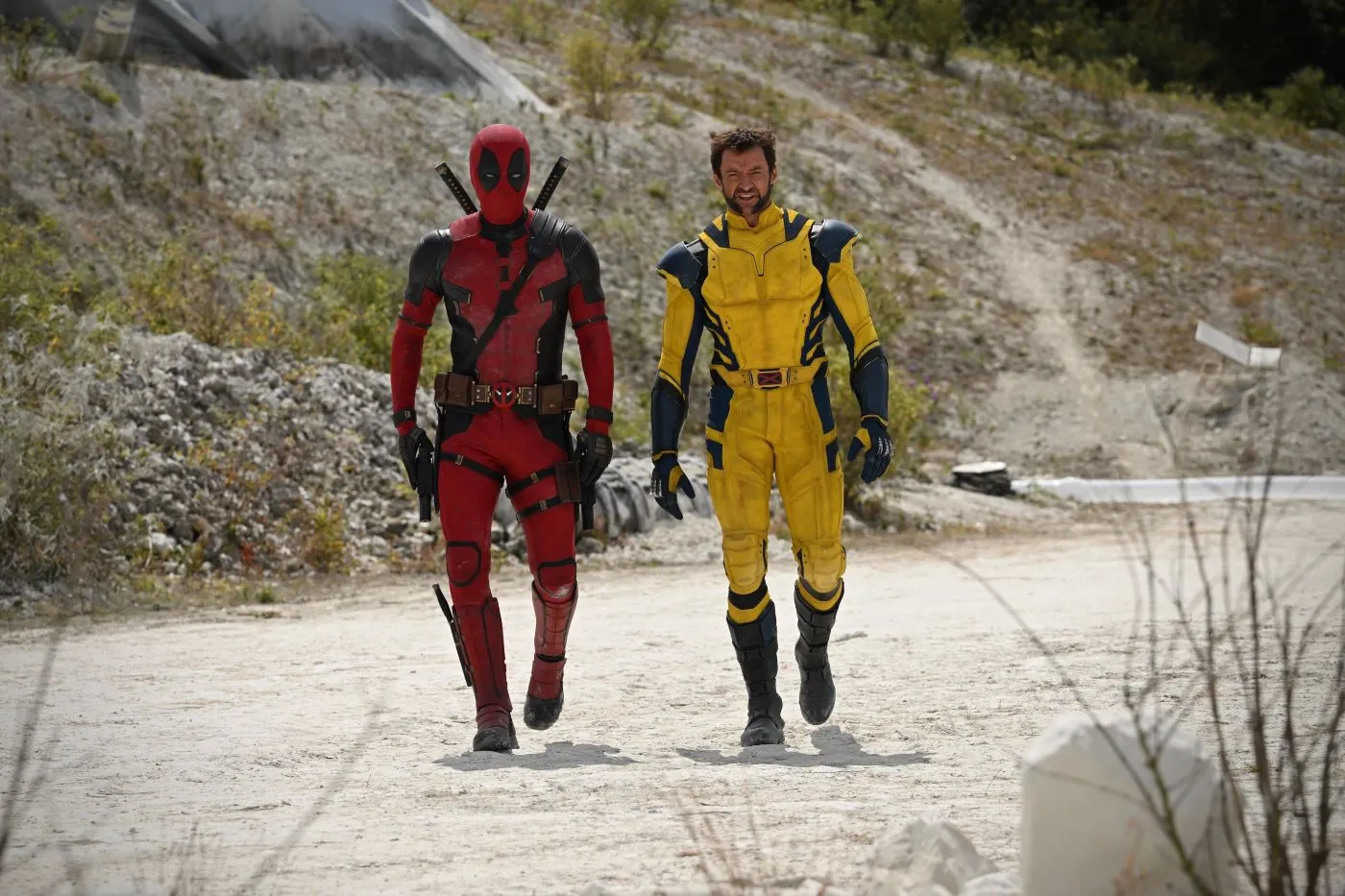 eine kahle, hügelige Wüstenlandschaft, mittig im Bild marschieren Deadpool im rot-schwarzen und Wolverine im gelb-blauen Anzug auf die Kamera zu
