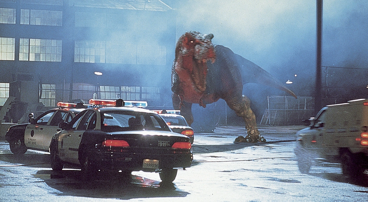 ein Parkplatz vor einer Lagerhalle bei Nacht, Polizeiautos fahren über die freie Fläche, vor ihnen baut sich ein Tyrannosaurus mit gefletschten Zähnen auf