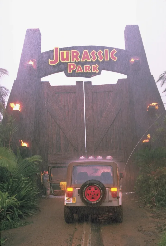 ein riesiges, hölzernes Eingangstor vor dem ein kleiner Wagen auf einer schienenartigen Spur steht, auf dem Torbogen in großen Lettern "JURASSIC PARK" geschrieben