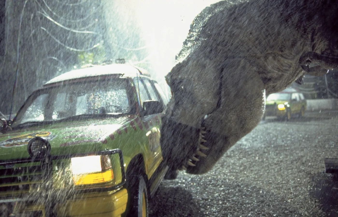 ein gelbes Auto steht auf matschigen Grund im Regen, ein Tyrannosaurus stößt mit seinem Kopf dagegen