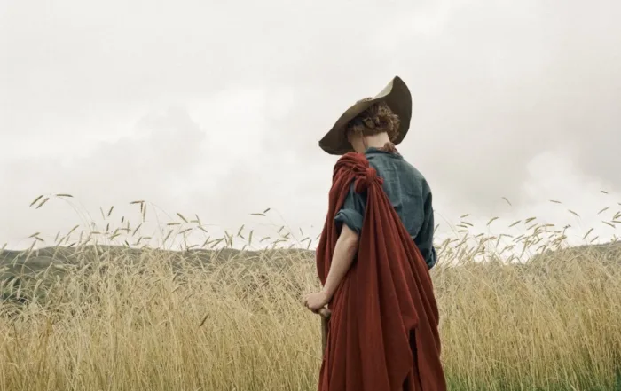 inmitten eines feldes steht eine kurzhaarige frau mit Hut, Gehstock, blauen Hemd und rotem übergehangenen Tuch