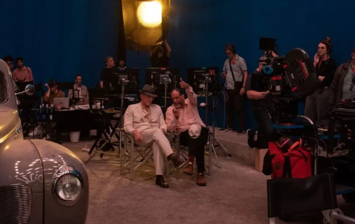 eine Filmcrew filmt in einem historischen Set, am linken Bildrand steht ein altes Auto, in der Mitte sitzen zwei Männern auf Regiestühlen und unterhalten sich, im Hintergrund arbeitet der Rest der Crew