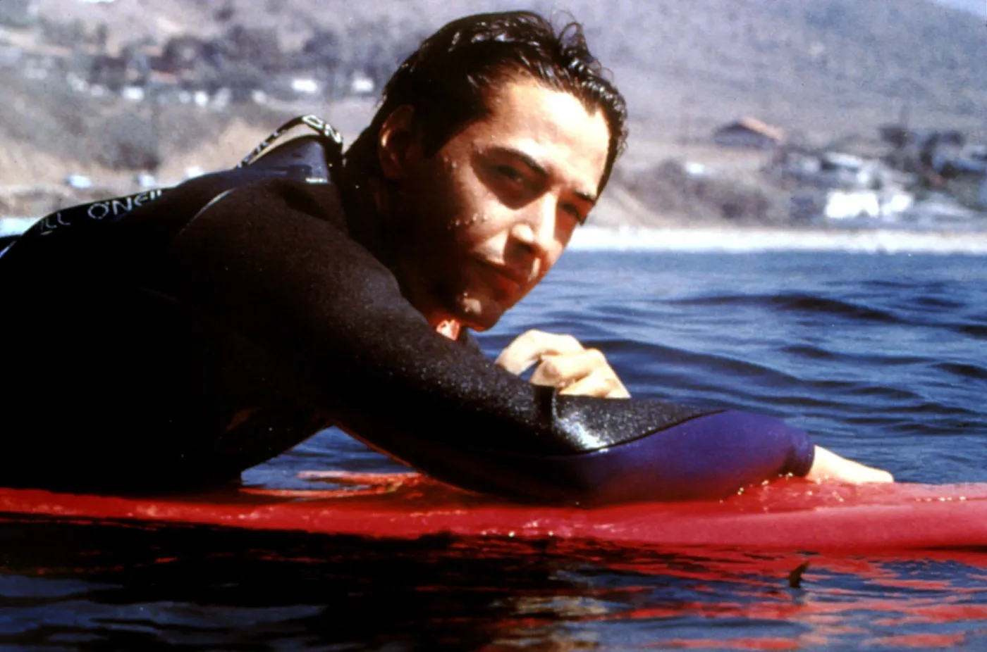 ein schwarzhaariger Surfer liegt auf einem roten Surfbrett im Wasser, im Hintergrund ist ein Küstenort zu sehen