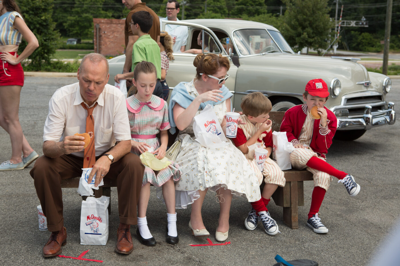 1950er Jahre, auf einer Holzbank sitzen ein Mann und eine Frau mit ihren drei Kindern, alle haben ein McDonalds-Produkt in der Hand und essen, der Mann schaut zu dem Burger in seiner Hand