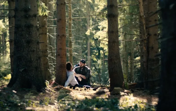 ein Wald bei Tag, an einem Baum gefesselt sitzt ein Mann im blauen Hemd, vor ihm hockt ein bärtiger Mann in dunkler Jacke