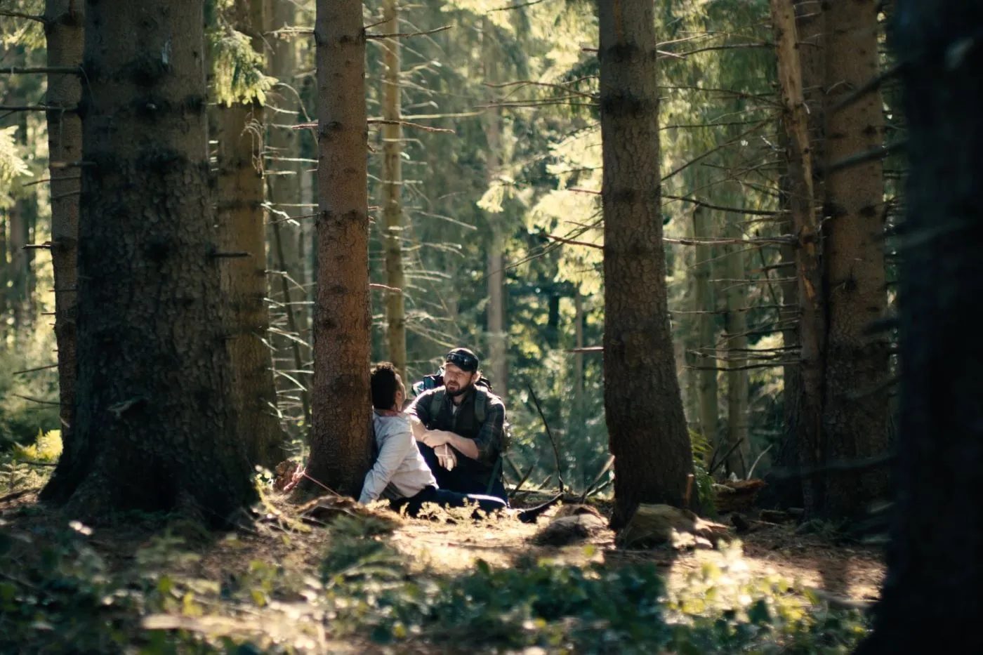 ein Wald bei Tag, an einem Baum gefesselt sitzt ein Mann im blauen Hemd, vor ihm hockt ein bärtiger Mann in dunkler Jacke