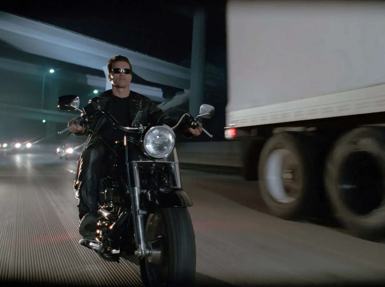 ein Mann mit verdunkelter Brille, Arnold Schwarzenegger als Terminator, fährt bei Nacht auf einem Motorrad auf der Autobahn