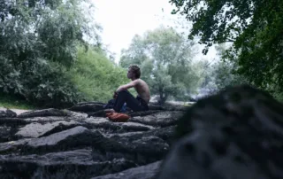 ein junger Mann sitzt oberkörperfrei auf einem von vielen kleinen Felsen, durch die sich Flusswasser schlängelt, um ihn herum sind die Bäume eines Waldes zu sehen