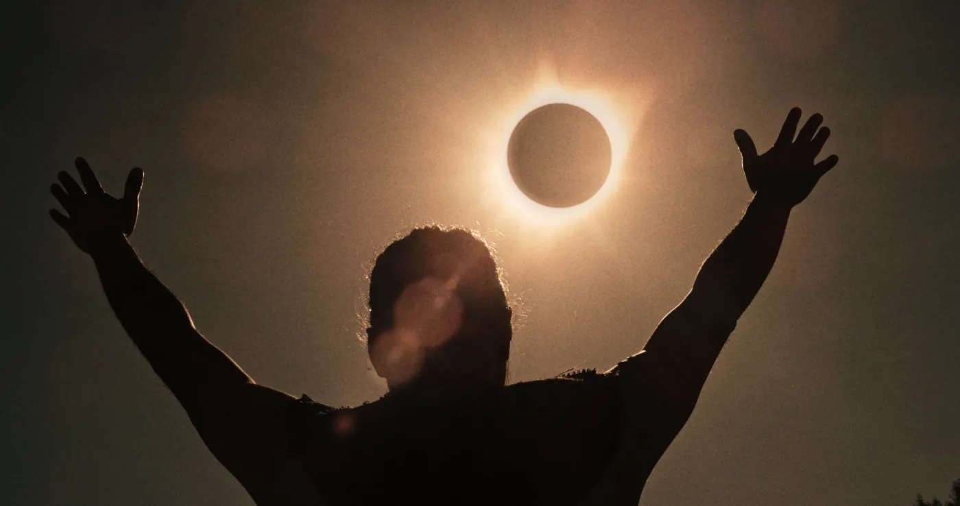 eine menschliche Gestalt reißt vor einer Sonnenfinsternis am Himmel beide Arme nach oben