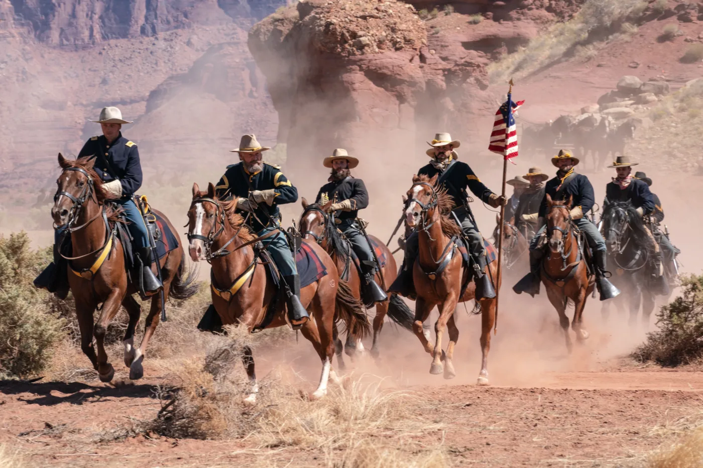 eine Männergruppe in dunklen Uniformen und mit hellbraunen Hüten reitet auf Pferden durch eine kahle, felsige Landschaft, einer der Männer trägt eine amerikanische Flagge bei sich