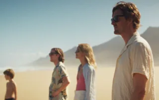 ein Strand, im Hintergrund bauen sich kleine Berge auf, eine Handvoll Menschen in kurzen Sachen und Sonnenbrille stehen am Strand und schauen aufs Meer