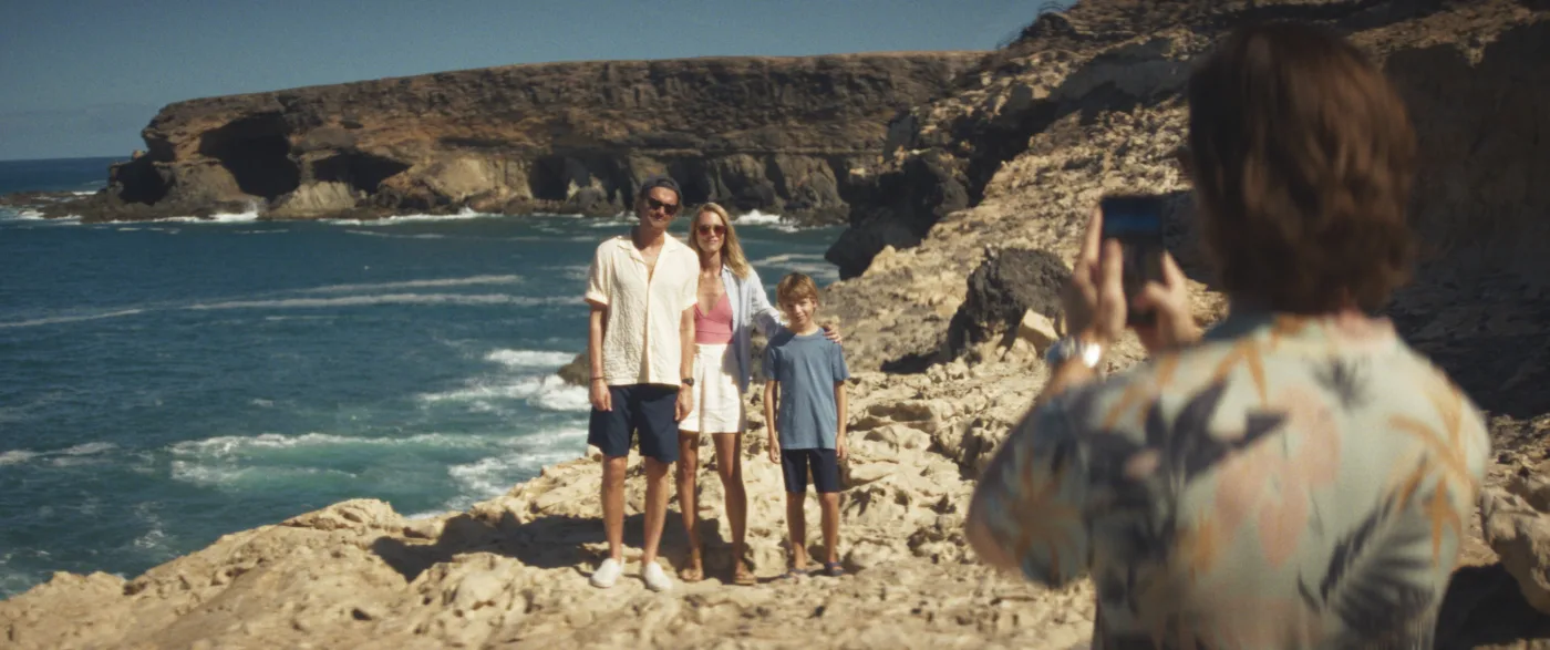 eine dreiköpfige Familie aus Vater, Mutter, Kind lässt sich vor einer sonnigen Küstenlandschaft fotografieren