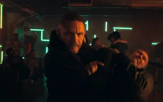 Tom Hardy als Undercover-Cop in einem Club, er hält einen Mann in der Mangel, während um ihn herum eine Schlägerei stattfindet
