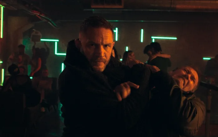 Tom Hardy als Undercover-Cop in einem Club, er hält einen Mann in der Mangel, während um ihn herum eine Schlägerei stattfindet