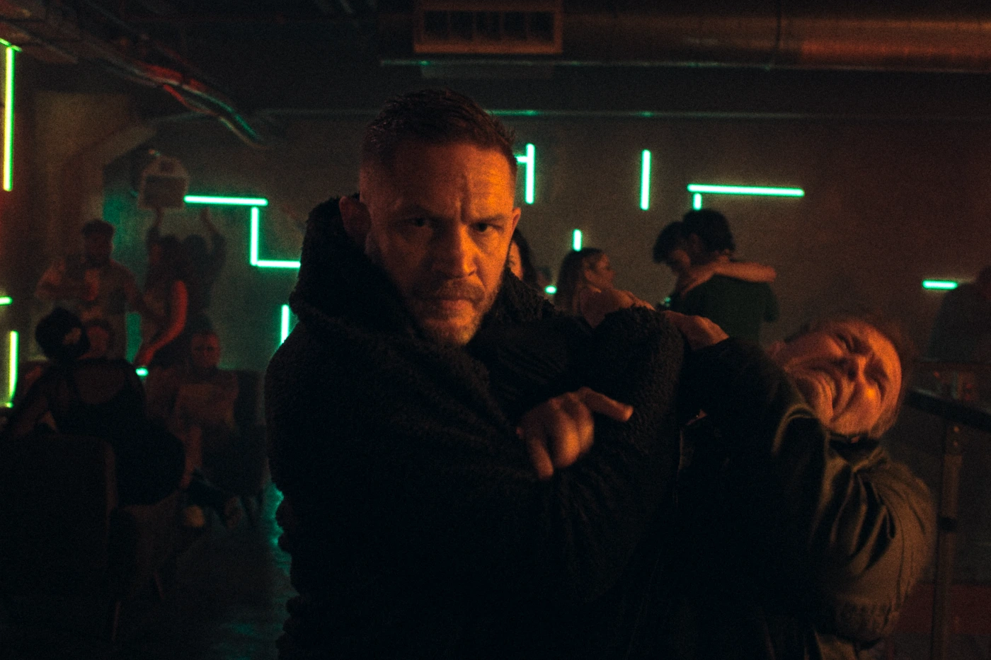 Tom Hardy als Undercover-Cop in einem Club, er hält einen Mann in der Mangel, während um ihn herum eine Schlägerei stattfindet