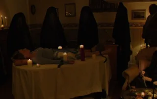 ein verdunkeltes Zimmer, in dessen Mitte ein Tisch mit weißer Tischdecke steht. Auf dem Tisch ist eine junge Frau gefesselt, Kerzen stehen neben ihr. Um den Tisch stehen Gestalten in dunklen Kutten, ganz rechts im Bild steht ein unmaskierter Mann.