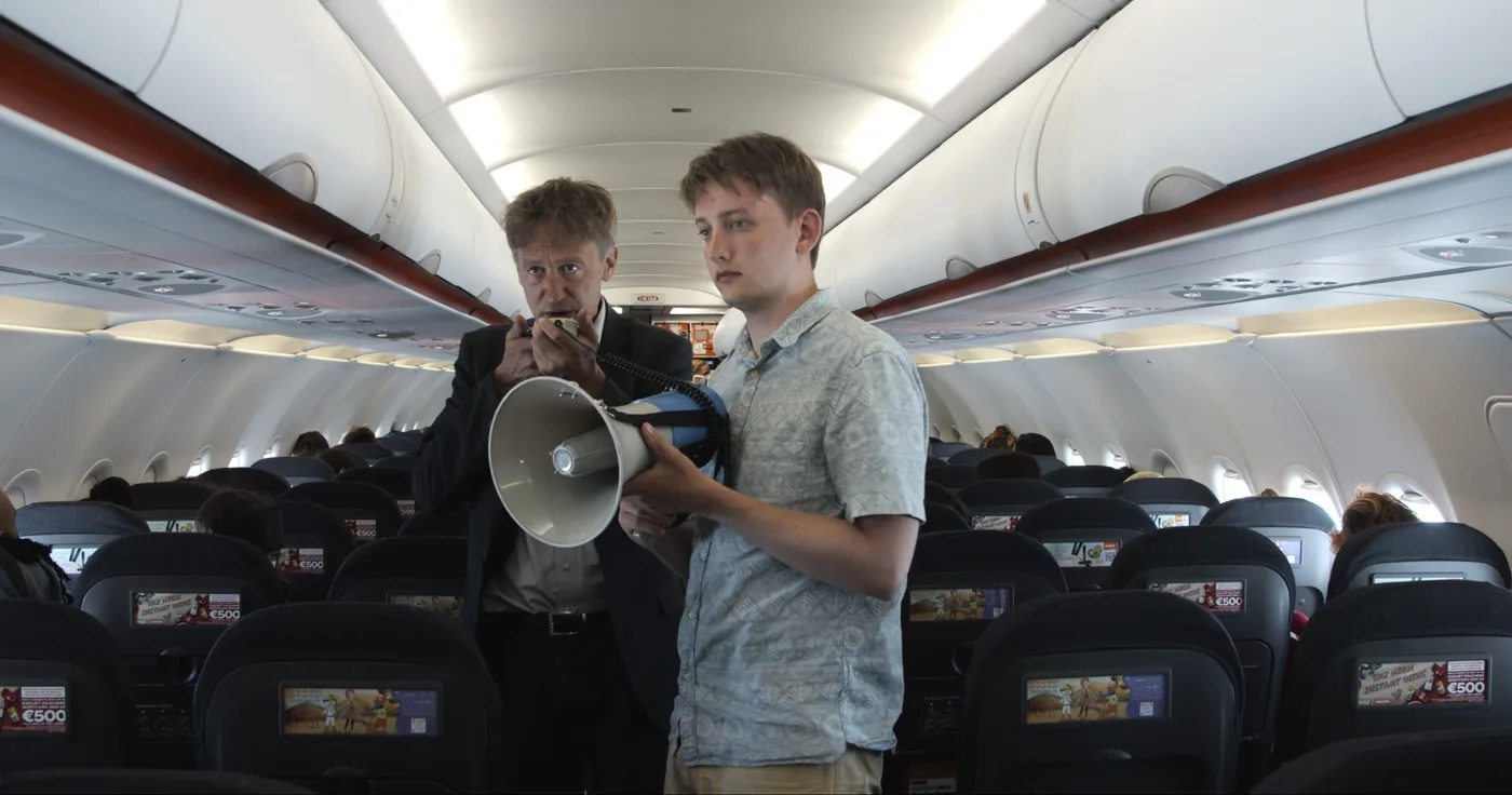 Jan Henrik Stahlberg als Mux im Gang eines Flugzeuges, ein junger Mann neben ihm, der ein Megafon hält