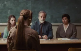 vor einer Tafel sitzen zwei Frauen und ein älterer Mann an einem Tisch, vor ihnen und mit dem Rücken zur Kamera sitzt eine Frau