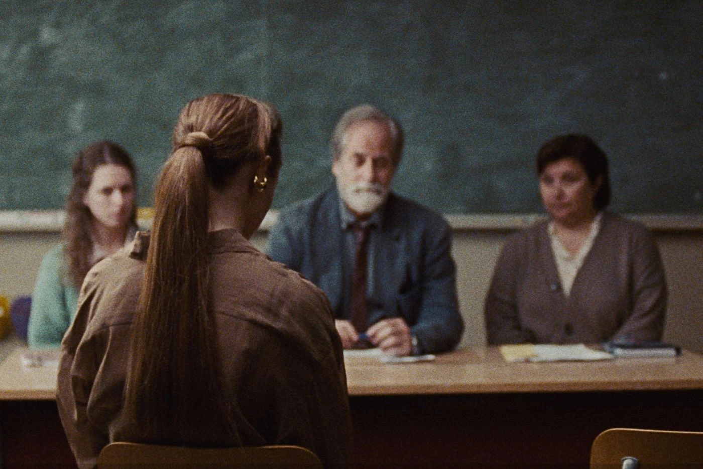vor einer Tafel sitzen zwei Frauen und ein älterer Mann an einem Tisch, vor ihnen und mit dem Rücken zur Kamera sitzt eine Frau