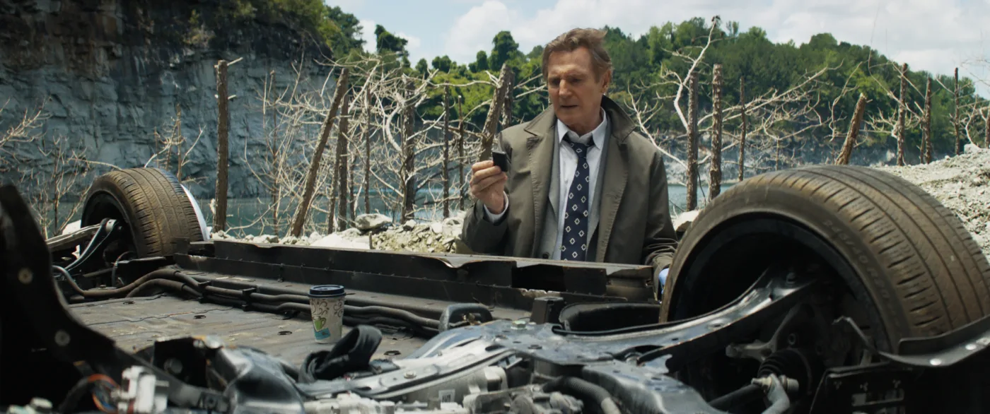 Liam Neeson als Frank Drebin Jr. vor einem verunfallten Autowrack, das auf dem Dach liegt