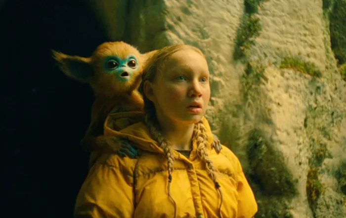 Helena Zengel als Yuri in gelber Regenjacke vor einem Felsen mit einem kleinen affenähnlichen Wesen auf der Schulter