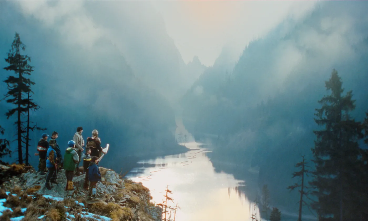 Ausblick in ein wolkenverhangenes Tal, durch welches ein Fluss führt, rechts im Bild auf einem Felsvorsprung eine Gruppe Jungen und ihr Vater