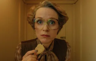 eine Frau mit dicker, großer Brille und einem Keks in der rechten Hand schaut intensiv in die Kamera