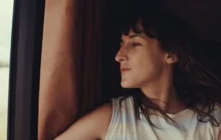 Eine Frau sitzt in einem Fahrzeug und schaut gedankenverloren aus dem Fenster. Ihr Oberkörper ist leicht zur Seite gedreht. Tageslicht fällt auf ihr Gesicht. Im Hintergrund ist ein brauner Vorhang zu sehen, der das Fenster teilweise bedeckt. Die Frau trägt ein ärmelloses, helles Oberteil mit sanften Streifen. Ihre Haare sind wellig und fallen ihr über die Schultern. (erstellt mit KI)