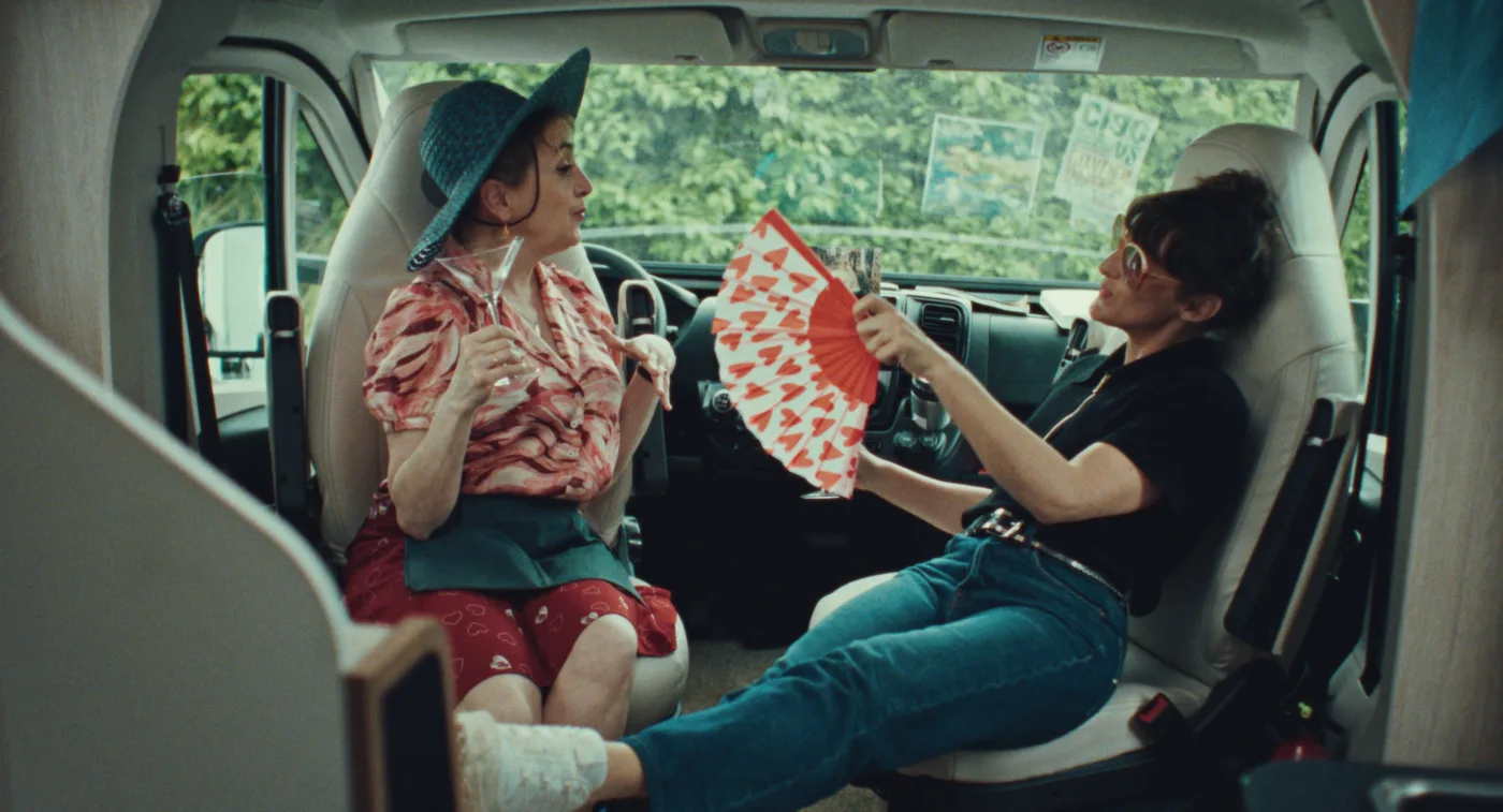 Zwei Frauen sitzen im vorderen Bereich eines Wohnmobils, auf den Fahrersitzen, die sich einander zugewandt sind. Die Frau links trägt einen gemusterten Hut und eine farbenfrohe Bluse. Sie hält ein Cocktailglas in der Hand. Die Frau rechts sitzt lässig mit ausgestreckten Beinen, trägt Sonnenbrille und Fächer mit rotem Muster in der Hand. Sie fächelt sich Luft zu. (erstellt mit KI)