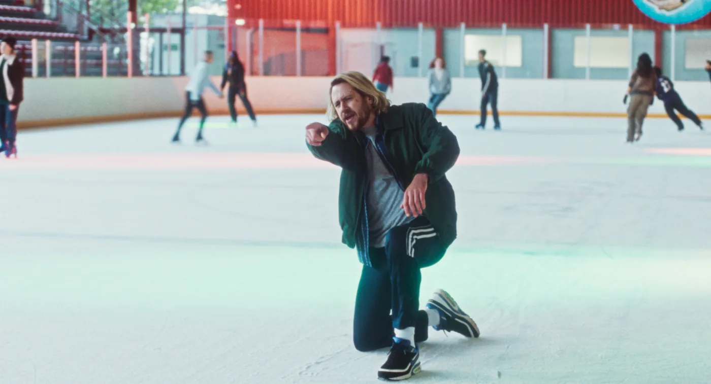 Ein Mann mit mittellangen, blonden Haaren kniet mitten auf einer Eislaufbahn. Er trägt eine grüne Jacke, dunkle Sporthose mit weißen Streifen und Turnschuhe mit Kufen.Mit entschlossenem Blick zeigt er mit dem Zeigefinger nach vorne. Im Hintergrund laufen weitere Personen über das Eis, teils verschwommen. Die Szene ist bunt beleuchtet, mit farbigen Lichtreflexen auf dem Eis. (erstellt mit KI)