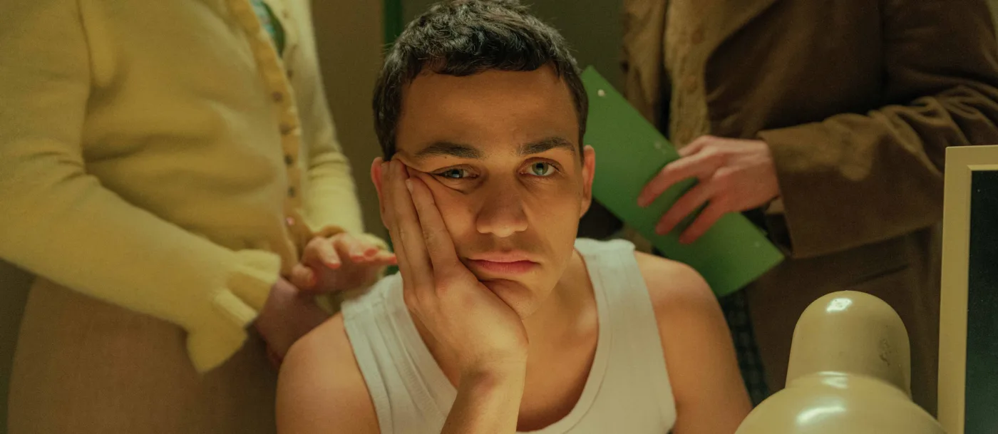 ein junger Mann im weißen Unterhemd an einem Tisch sitzend, den Kopf auf den aufgestützten rechten Arm gestützt