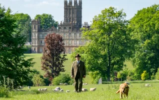 ein Mann im braunen Anzug und Hut auf einer Wiese, im Hintergrund liegt Downton Abbey