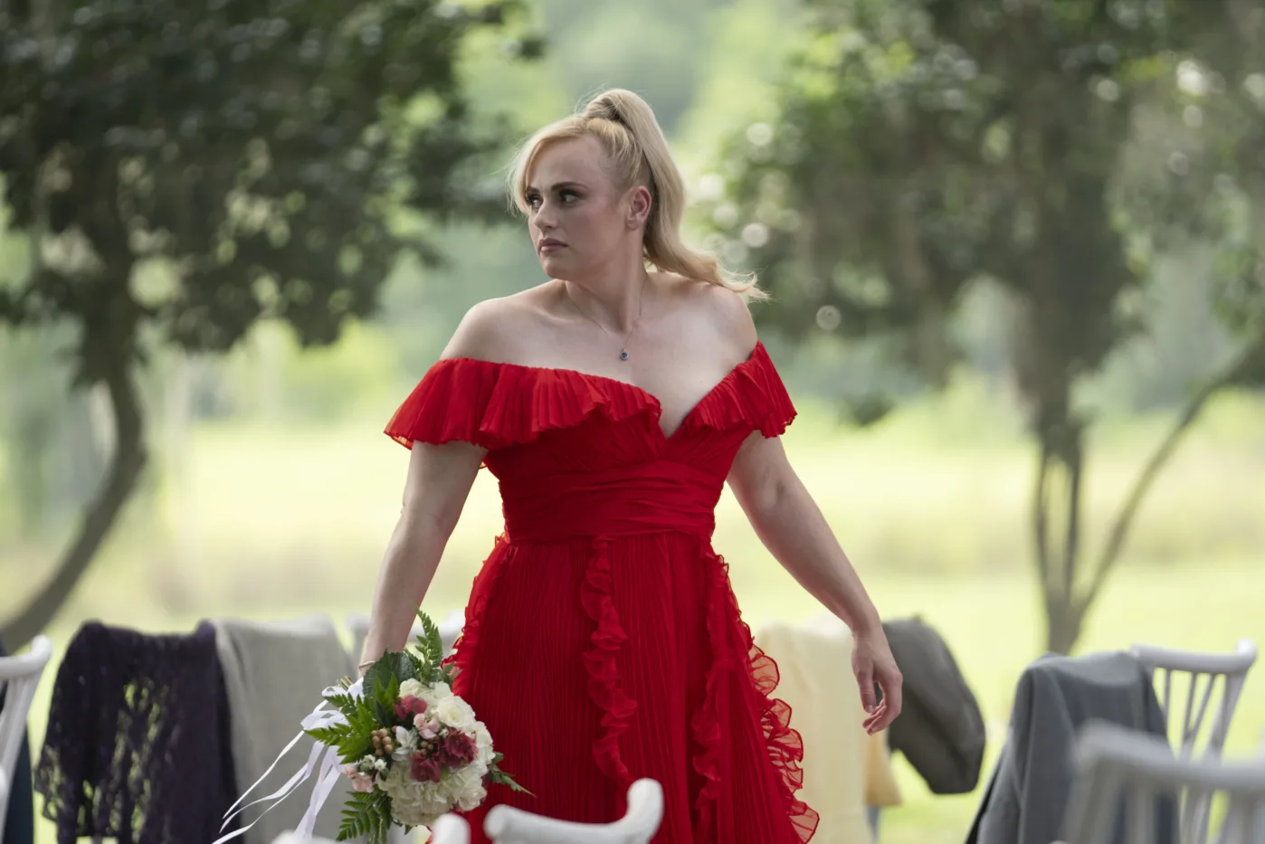 eine blonde Frau in rotem Kleid mit einem Blumenstrauß in der rechten Hand und aufgewühltem Gesichtsausdruck