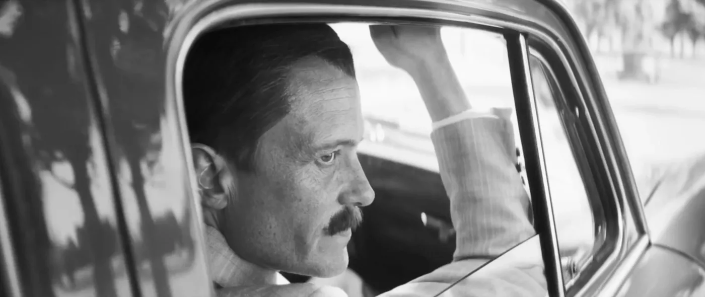 Ein Mann sitzt auf dem Beifahrersitz eines Autos und hält sich mit der rechten Hand fest. Er trägt einen hellen Anzug mit feinen Streifen. Das Auto hat eine abgerundete Fensteröffnung und eine glänzende Oberfläche. Im Hintergrund sind unscharfe Bäume und ein Gehweg zu erkennen. [erstellt mit KI]