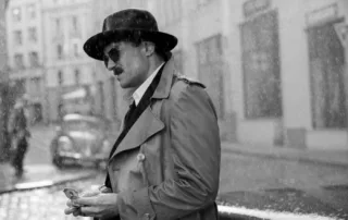 Ein Mann trägt einen dunklen Hut und einen hellen Trenchcoat mit doppelreihiger Knopfleiste. Er steht auf einem Kopfsteinpflaster vor einem Gebäude mit großen Fenstern. In seiner rechten Hand hält er eine kleine Schachtel oder ein ähnliches Objekt. Im Hintergrund sind unscharf weitere Gebäude und ein geparktes Auto zu erkennen. [erstellt mit KI]