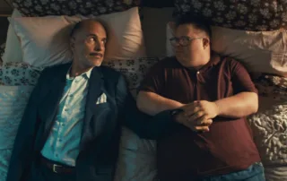 Zwei Männer liegen nebeneinander auf einem Bett mit gemusterten Bettlaken und mehreren Kissen. Der Mann links trägt einen dunklen Anzug mit weißem Hemd und einem Einstecktuch, der Mann rechts ein dunkelrotes Poloshirt und Jeans. Beide haben die Arme entspannt auf dem Bauch verschränkt. [erstellt mit KI]