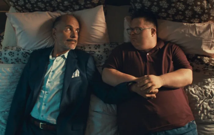 Zwei Männer liegen nebeneinander auf einem Bett mit gemusterten Bettlaken und mehreren Kissen. Der Mann links trägt einen dunklen Anzug mit weißem Hemd und einem Einstecktuch, der Mann rechts ein dunkelrotes Poloshirt und Jeans. Beide haben die Arme entspannt auf dem Bauch verschränkt. [erstellt mit KI]