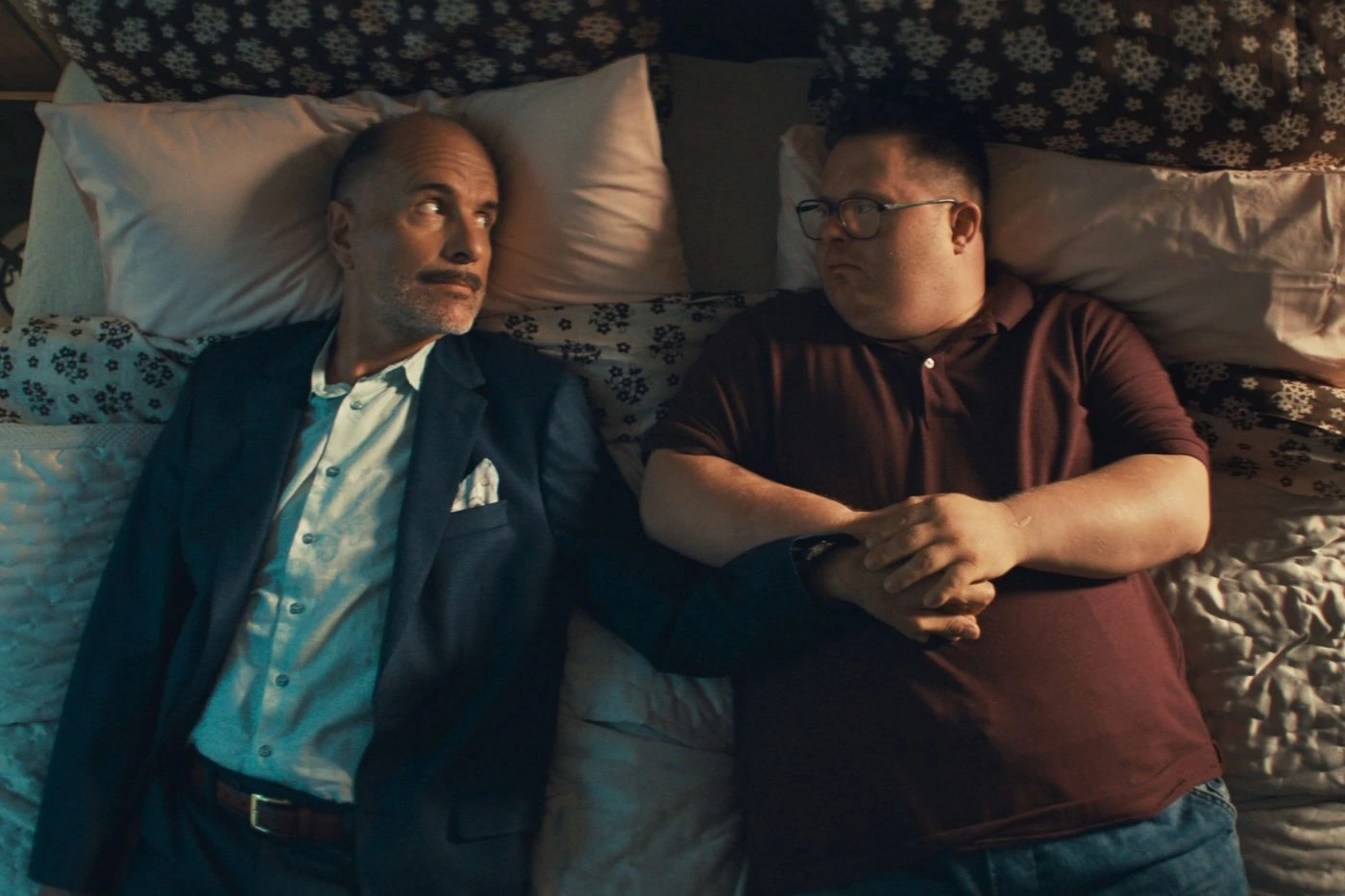 Zwei Männer liegen nebeneinander auf einem Bett mit gemusterten Bettlaken und mehreren Kissen. Der Mann links trägt einen dunklen Anzug mit weißem Hemd und einem Einstecktuch, der Mann rechts ein dunkelrotes Poloshirt und Jeans. Beide haben die Arme entspannt auf dem Bauch verschränkt. [erstellt mit KI]