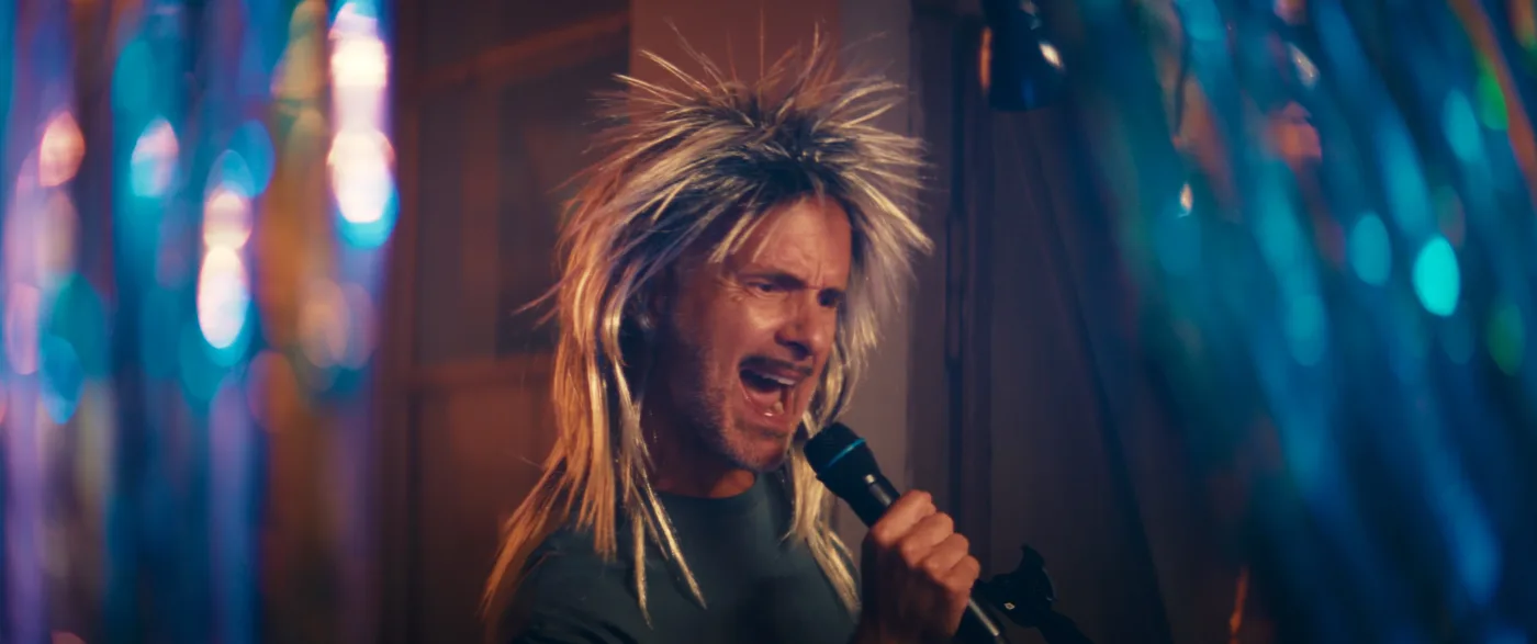 Eine Person mit einem schwarzen T-Shirt hält ein Mikrofon in der rechten Hand. Die Haare sind lang, blond und stachelig frisiert. Im Hintergrund sind unscharfe bunte Lichter zu sehen, die eine festliche Atmosphäre erzeugen [erstellt mit KI]