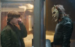 Ein junger Mann mit braunen, schulterlangen Haaren trägt eine grüne Jacke, steht in einem Telefonzellenhäuschen und hält einen Telefonhörer an sein Ohr. Draußen, ihm gegenüber, steht eine Person mit einer weißen, tierähnlichen Maske, die spitze Ohren und eine lange Schnauze zeigt. Die maskierte Person trägt dunkle Kleidung und hat lange, zerzauste Haare. [erstellt mit KI]
