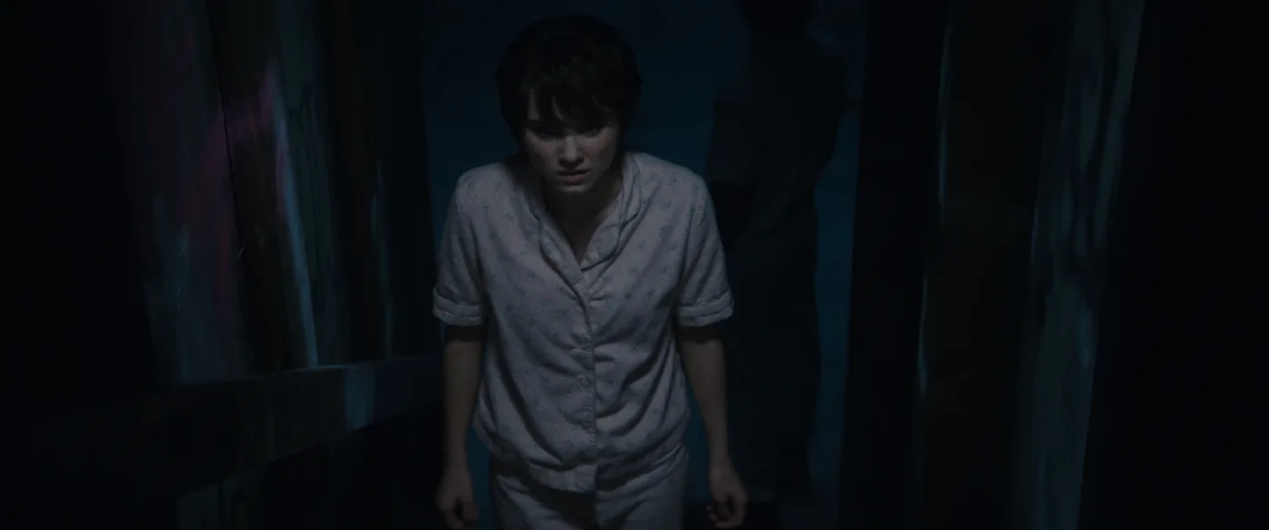 Eine Person in weißer, kurzärmeliger Kleidung steht in einem dunklen, engen Flur. Die Arme hängen seitlich herab, und die Person blickt nach vorne. Die Umgebung ist schwach beleuchtet, mit dunklen Wänden und einem schmalen Durchgang. Im Hintergrund sieht man eine Gestalt im Dunkeln stehen. [erstellt mit KI]