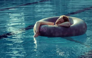 Eine junge Person liegt entspannt in einem grauen Schwimmreifen in einem hellblauen Swimmingpool. Sie ruht ihren Kopf und einen Arm auf dem Reifen und blickt ruhig ins Wasser. Auf dem Reifen ist in rosa Buchstaben das Wort „BAD“ zu lesen. [KI generiert]