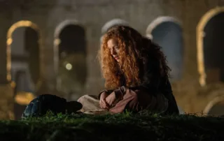 Ein Mädchen mit langen, lockigen Haaren sitzt auf dem Boden, die Arme auf den Knien verschränkt. Sie trägt dunkle Kleidung und sitzt auf einer grasbewachsenen Fläche. Im Hintergrund sind unscharf große, steinerne Bögen zu erkennen, die an eine historische Ruine erinnern. [erstellt mit KI]